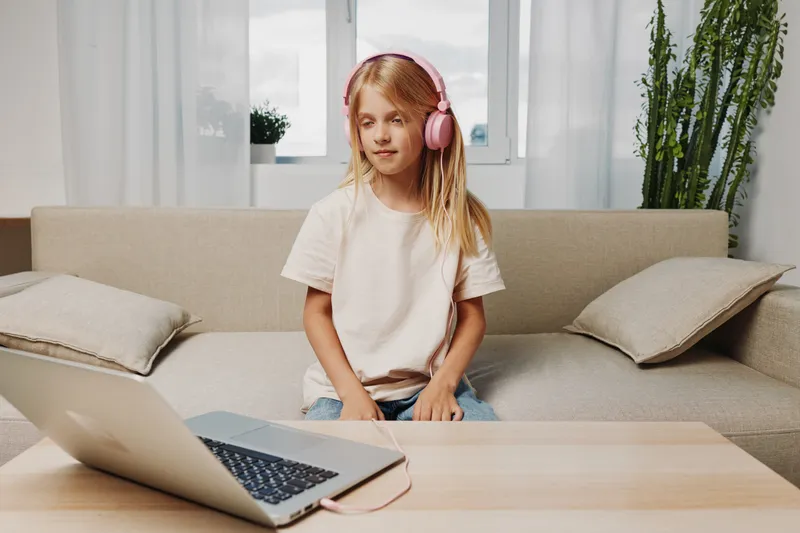 Young girl with headphones sitting on sofa using laptop at home, casual atmosphere with natural light and cozy interior behind her.