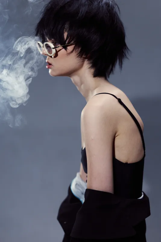 Young woman with short black hair wearing round glasses smoking against gray background