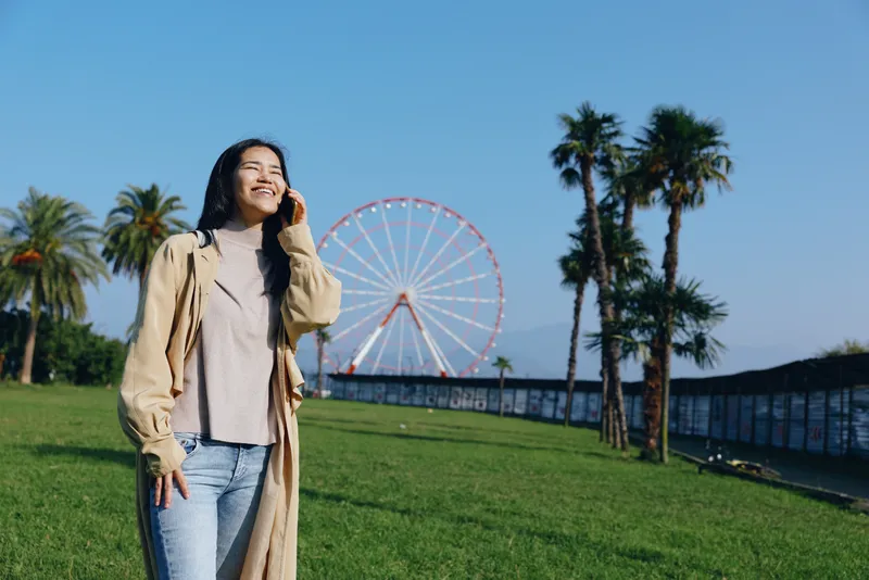 Woman in a beige coat strolls through a sunny park with a ferris wheel in the background, palm trees and green grass creating a joyful outdoor scene