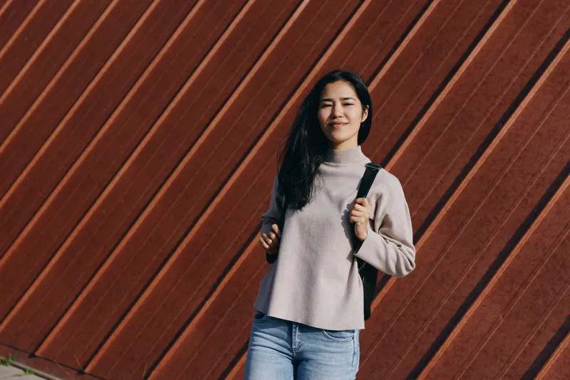 Casual outdoor scene featuring a woman with a backpack, wearing a sweater and denim, standing against a diagonal wooden wall. Bright, relaxed mood with urban style and fashion appeal for lifestyle,