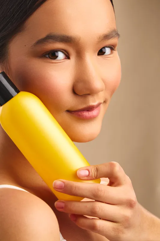 Asian woman holds a yellow bottle to her ear, smiling softly in a warm indoor setting, creating a relaxed and confident casual office vibe.