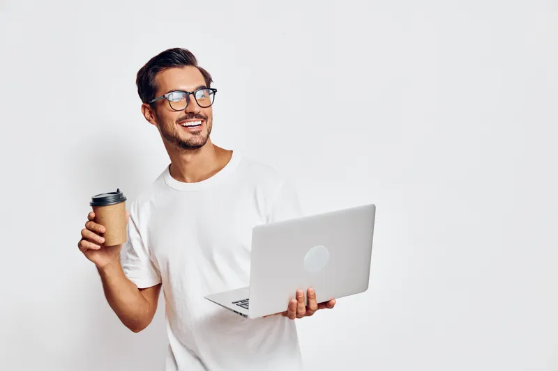 Smiling man holding a coffee cup while using a laptop, looking happy and relaxed against a plain solid background. People lifestyle concept.