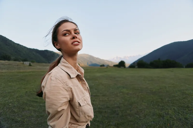Young woman with long brunette hair stands in an open field surrounded by mountains during sunset, wearing a beige jacket and looking confidently into the distance.
