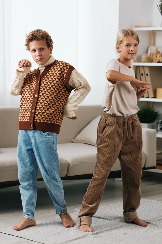 Two playful children pose in a bright living room, wearing cozy knitwear and casual denim, captured in soft natural light with a calm, homey atmosphere.