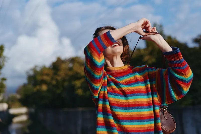 woman sweater colorful photography camera outdoor young woman in rainbow striped knitwear taking a candid photo, smiling and looking up under blue sky with sunlight and trees in background