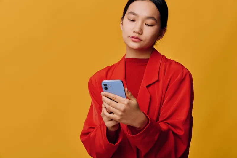 Young woman wearing red jacket using smartphone on yellow background. Female holding phone with focus on screen for communication, technology, social media, and connection concept.