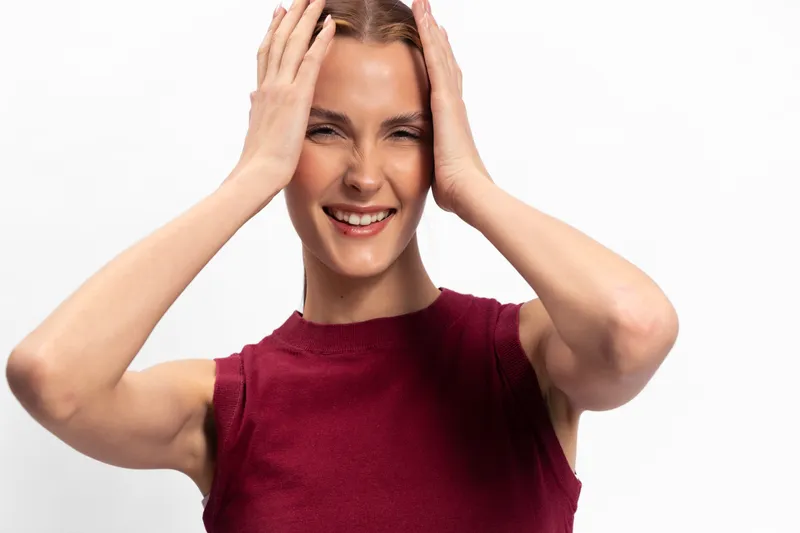 Smiling young woman with hands on head wears sleeveless burgundy top, isolated on white. Concept for happiness, confidence, positive emotions, and casual lifestyle portrait.