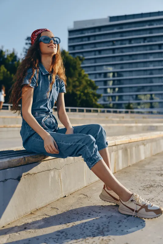 Young woman wearing denim jumpsuit and sunglasses sitting outdoors on a sunny day with modern architecture and green trees in the background.