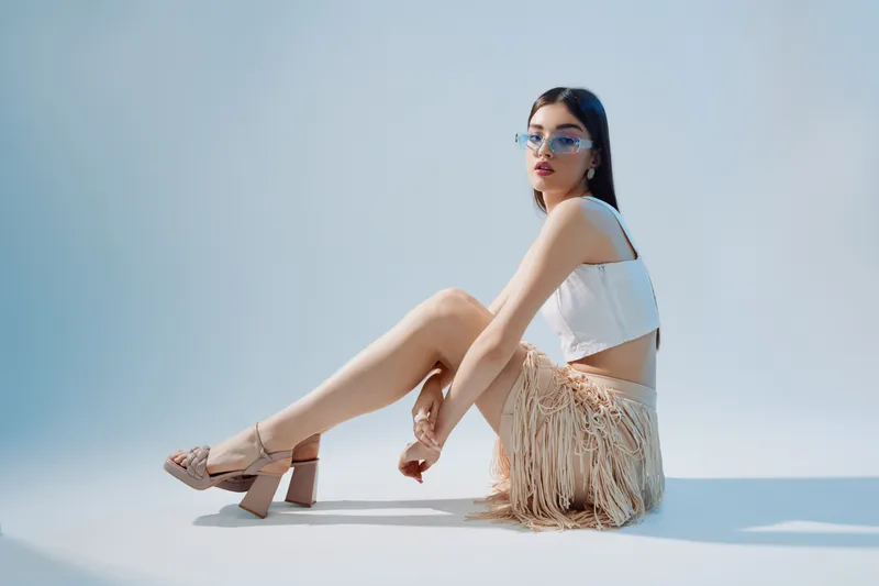 Young woman in fashionable outfit and sunglasses sits on floor against light background. Concept of woman beauty fashion and modern style in studio lighting.