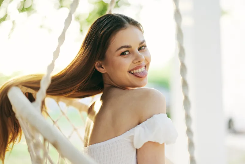 Young woman with long hair smiling playfully on a swing, showcasing carefree summer vibes with a warm backdrop and natural lighting