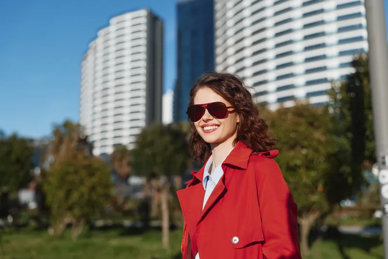 Young woman in a red coat smiles in bright sunlight against a modern cityscape, wearing sunglasses and enjoying a candid outdoor moment.