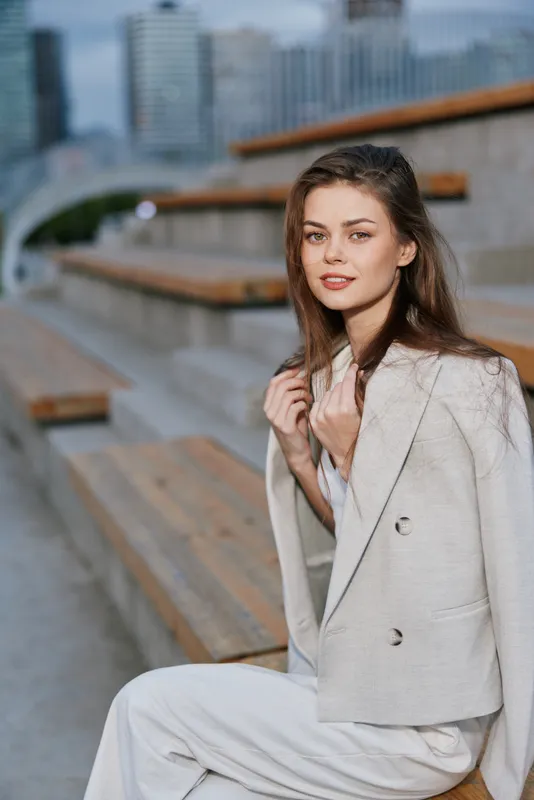 Young woman in light blazer sitting on urban stairs with confident expression and natural makeup in soft daylight fashion portrait