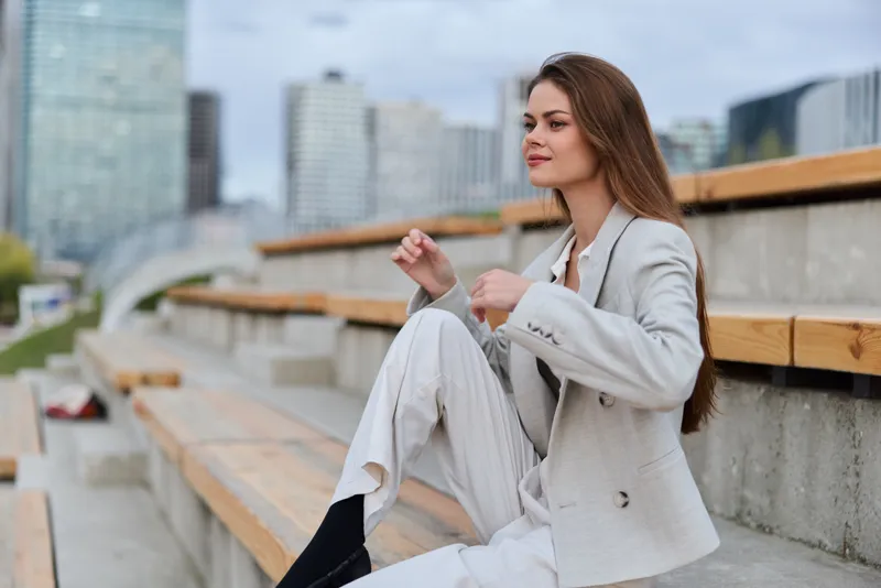 Young confident woman in light suit sitting outdoors on urban benches with cityscape background, showcasing thoughtful expression and soft natural tones in business style