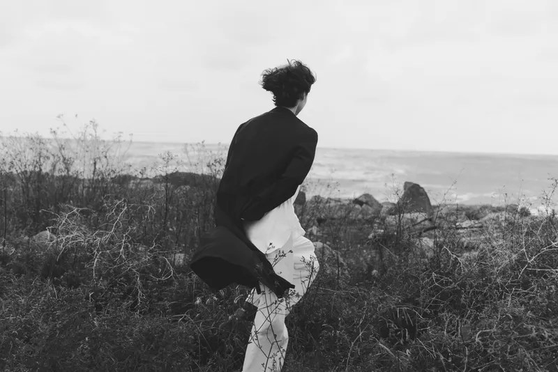 A windswept silhouette of a woman standing by a rugged coastline, coat flaring in the breeze, captured in moody grayscale. A solitary moment of calm and motion, emphasizing natural textures, sea