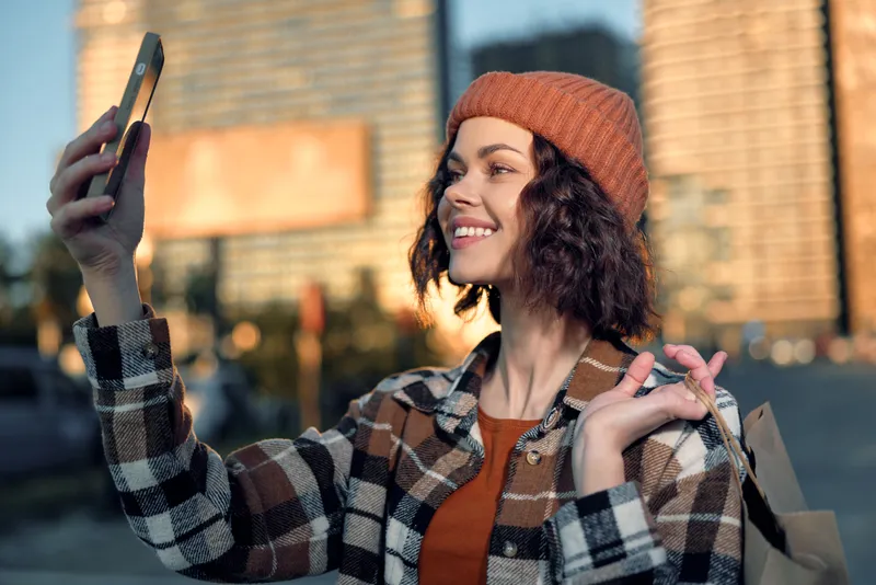 Woman, selfie, smile, shopping, city, smartphone capturing golden hour glow while holding a bag, candid authenticity and mindful living mood, emotional storytelling portrait of modern urban lifestyle