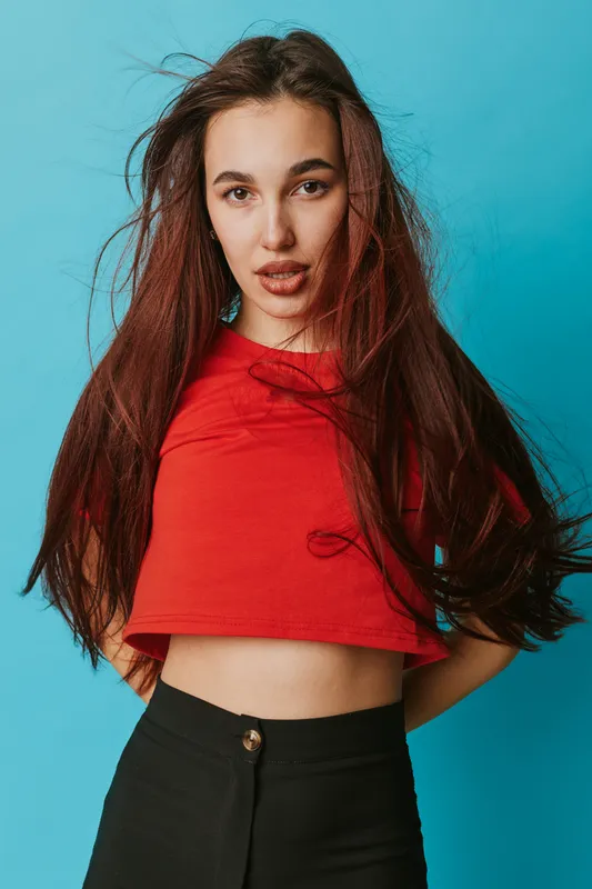 Young woman with long brown hair wearing red cropped top and black pants posing against blue background. Casual style, confident expression, and natural makeup for fashion portrait.