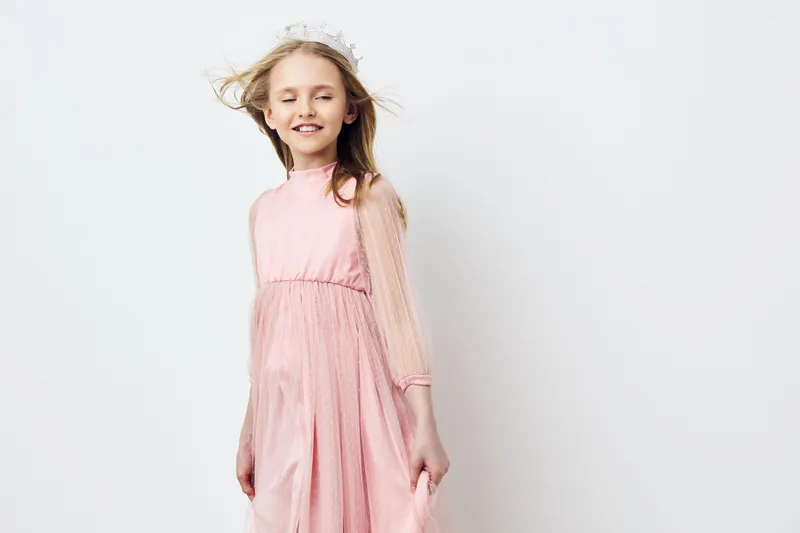 A joyful young girl in a light pink dress smiles while standing against a clean white background, showcasing a carefree and playful atmosphere