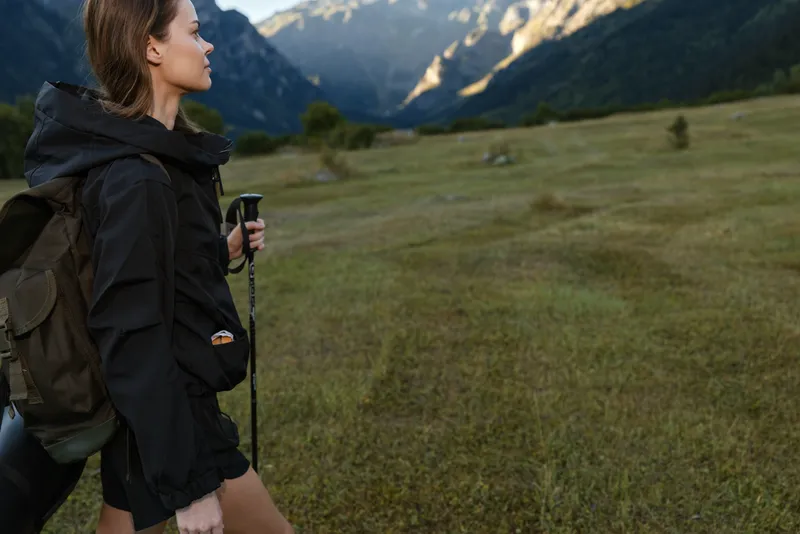 A female hiker with a backpack stands outdoors in a mountain meadow, holding a trekking pole. She looks ahead with calm focus, enjoying nature in a rugged alpine landscape.