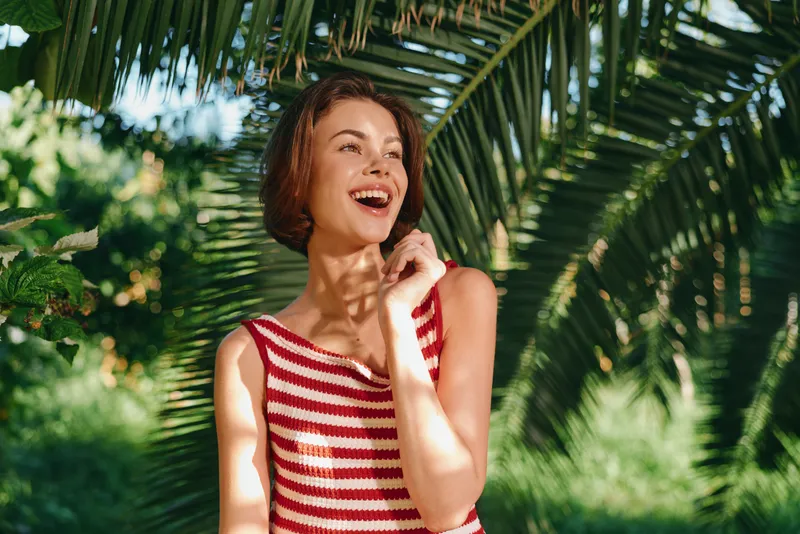 A cheerful woman in a striped red and white top smiles brightly in a sunny outdoor setting, framed by lush tropical palms and vibrant green foliage, creating a warm, inviting mood and a lively,