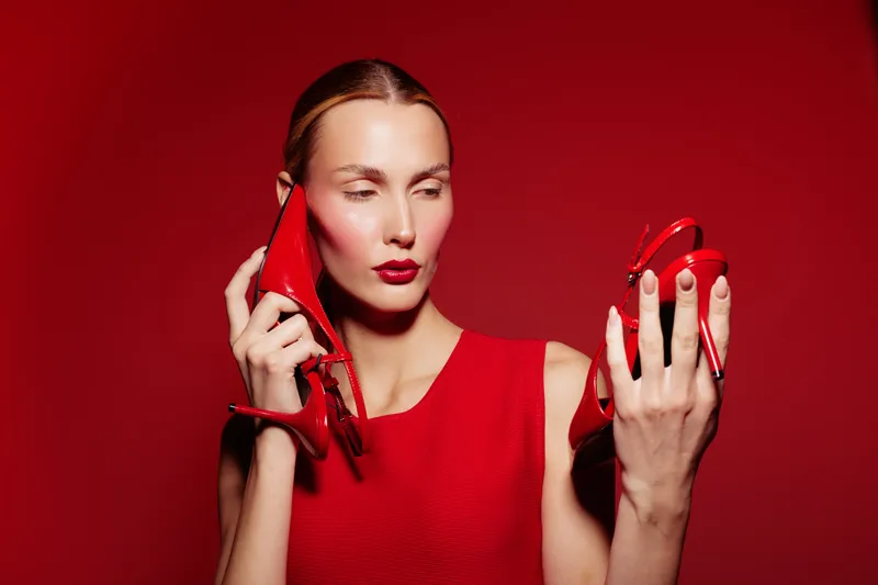 Young woman in red dress holds red high heels, one near her ear like a phone, against red background. Concept of fashion, style, creativity, and bold color contrast.