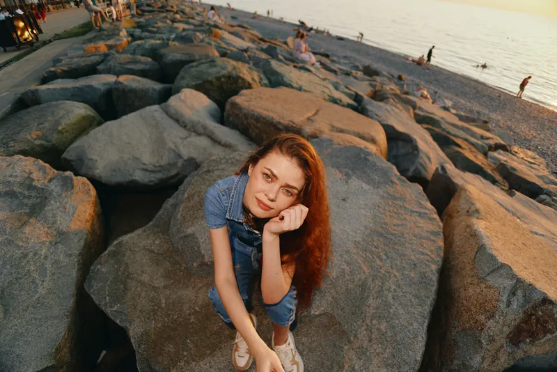 young woman with red hair sitting thoughtfully on large beach rocks in warm sunset light, casual denim outfit, peaceful coastal scene