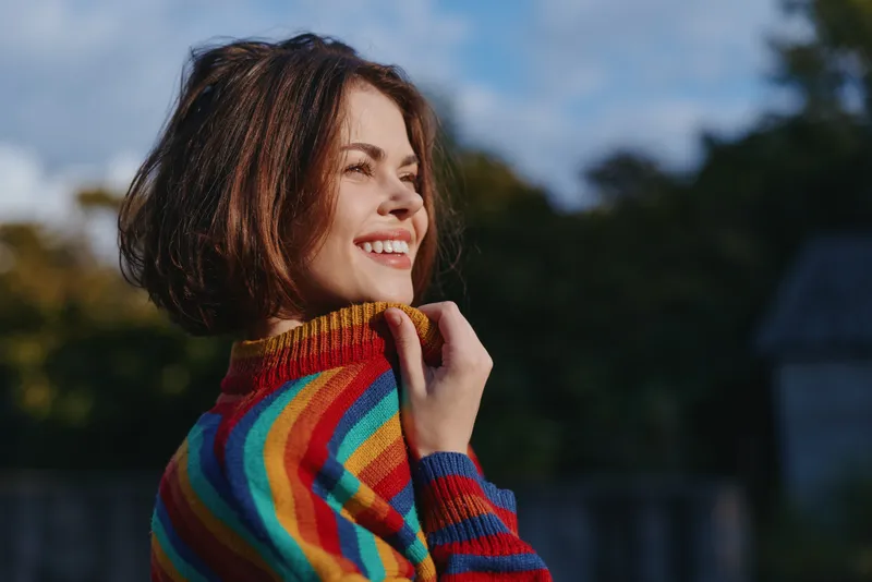 Woman smile portrait in colorful sweater outdoors, happy young woman with short hair looking away in natural light, candid expression and casual cozy fashion.