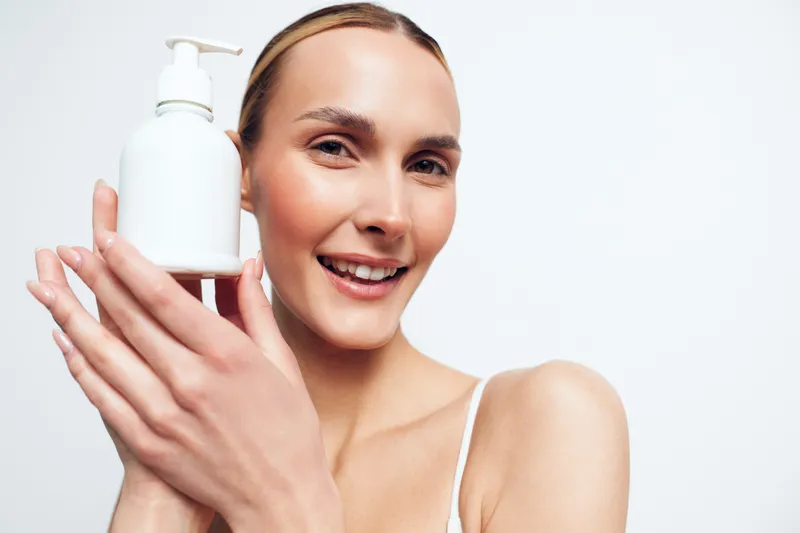 Smiling woman applying skincare, holding pump bottle near face, smooth skin and natural glow, minimal white background, clean beauty hygiene, close up, soft light