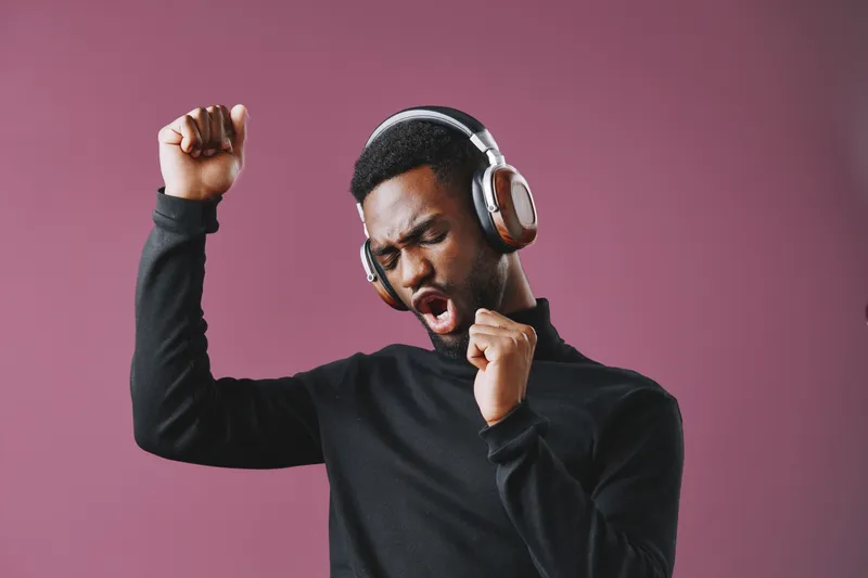 Young Black man enjoying music with headphones, expressing joy and excitement, wearing a black turtleneck against a soft pink background, conveying a modern and youthful vibe