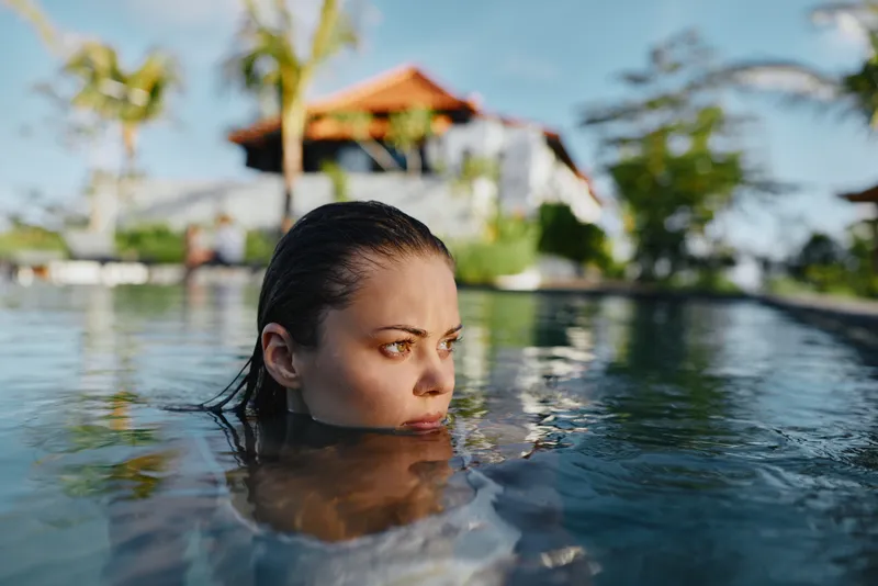 Relaxed woman in a sunlit pool, head above water, calm expression, gentle breeze, tropical backdrop with palm trees and a villa, vacation mood, casual swimwear, candid lifestyle