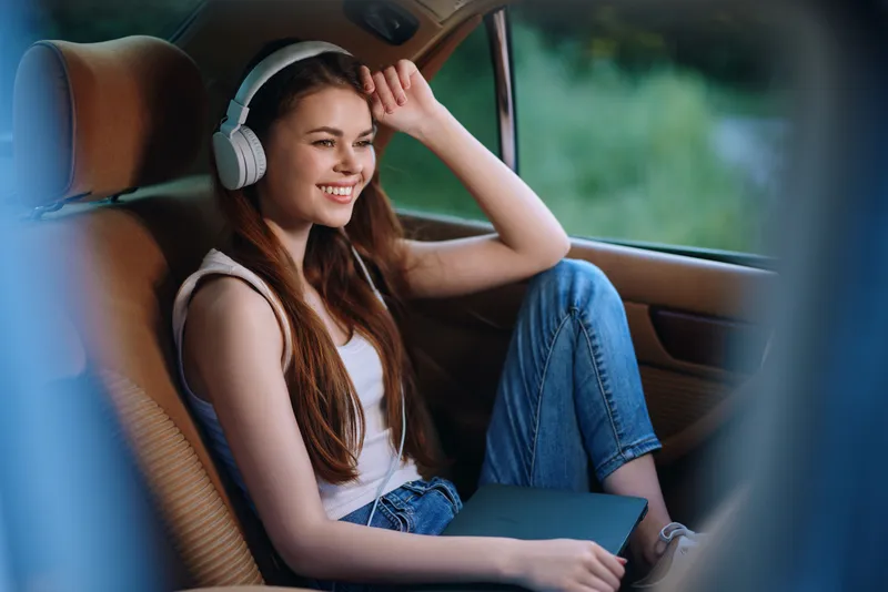 Happy young woman relaxing in a car with headphones, enjoying music while holding a laptop, casual attire, bright green background, youthful vibe.
