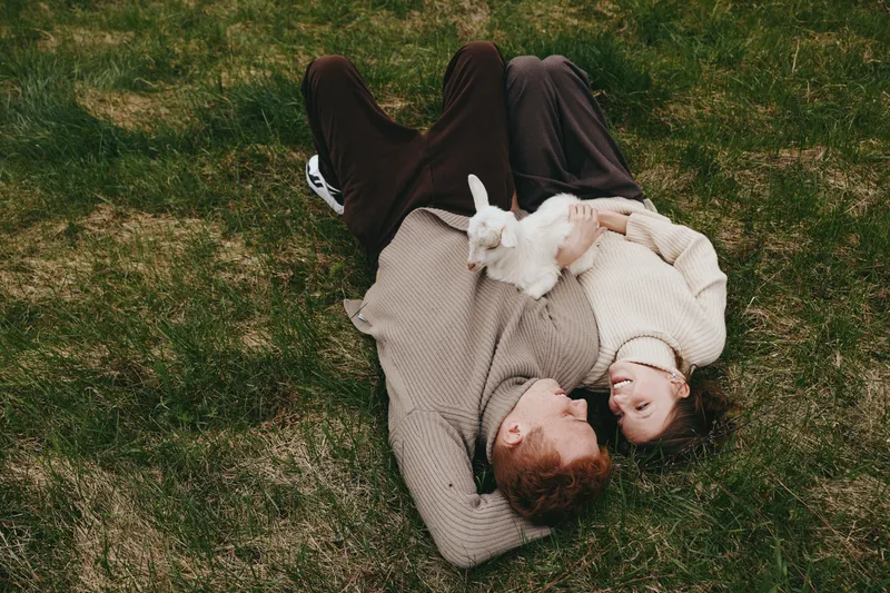 Relaxed couple lying on a grassy lawn with a playful goat kid resting between them, their warm embrace and soft smiles capture a candid outdoor family moment in peaceful nature.