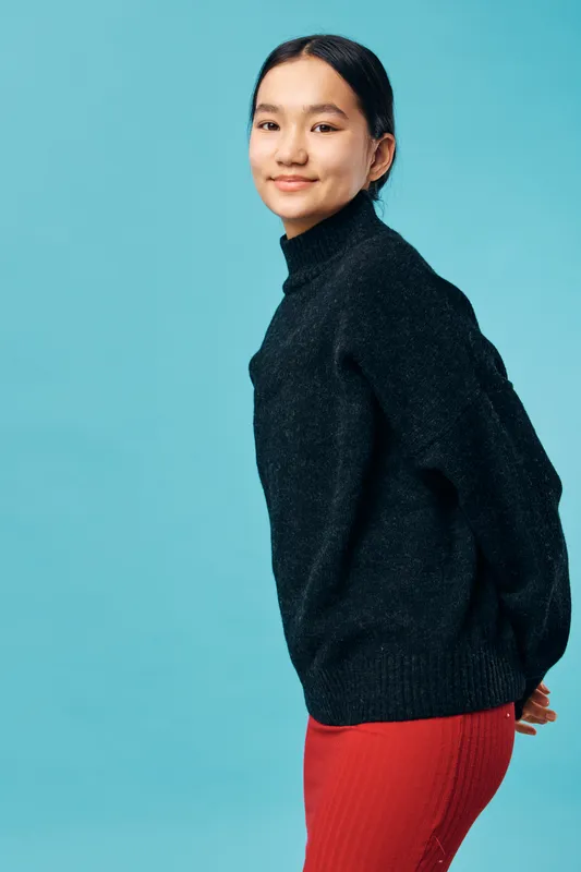 Young woman posing in black sweater and red skirt against blue background, smiling gently with casual posture and natural makeup, studio portrait of confident female model.