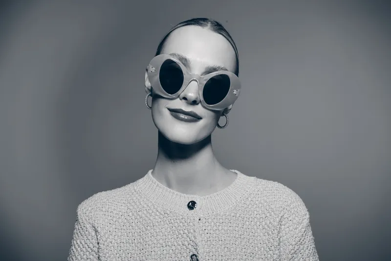 Young white woman with light skin wearing large round sunglasses and a knitted sweater looks confidently at camera in studio. Fashion and style concept.