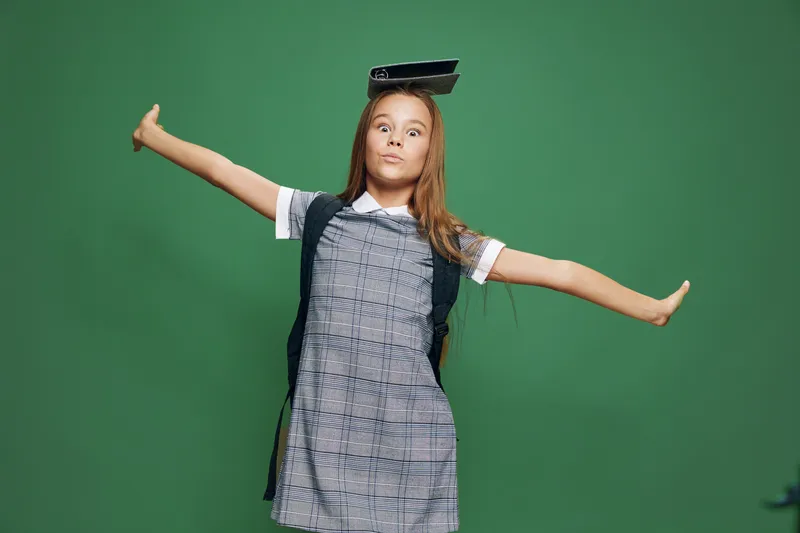 Girl child balancing book on head with arms outstretched for fun smiling in dress standing on green background in studio playful confident pose.