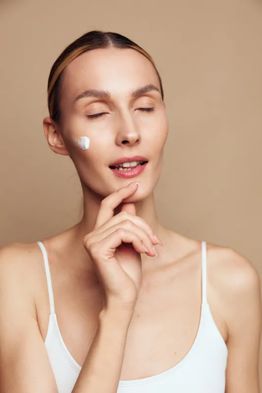 Young woman with closed eyes applies face cream on cheek, showing natural skin care routine, minimal makeup, and calm expression against beige background.