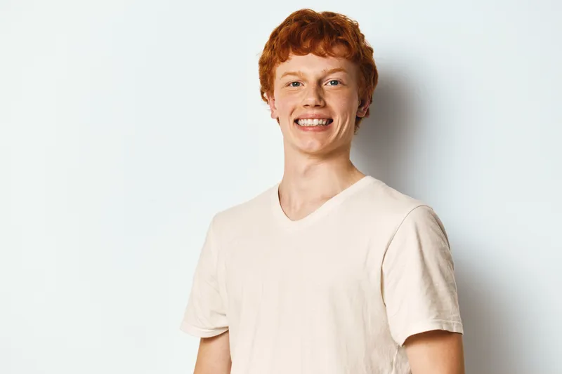 Young man with red hair and freckles smiling wearing light t shirt on white background. Portrait of cheerful casual guy in studio with relaxed posture and friendly expression.