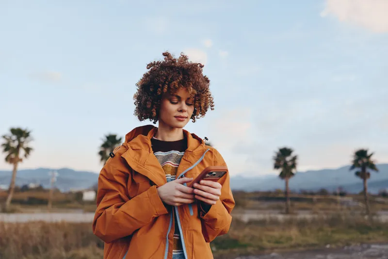 Woman lifestyle outdoors, holding coffee cup, wearing orange jacket, curly hair, gentle smile, warm sunset glow, palm trees, distant mountains, relaxed everyday walk in open countryside
