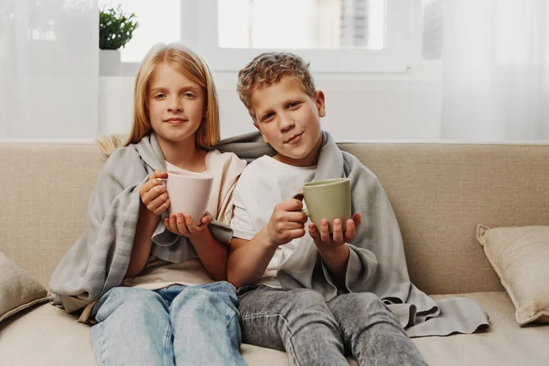 Two children wrapped in blankets sit on a sofa holding warm mugs, enjoying a cozy indoor moment with soft natural light from the window behind them.
