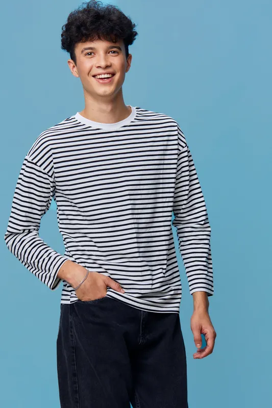 smiling young man wearing striped long sleeve shirt and black pants standing relaxed on blue background with casual pose and curly hair, joyful expression