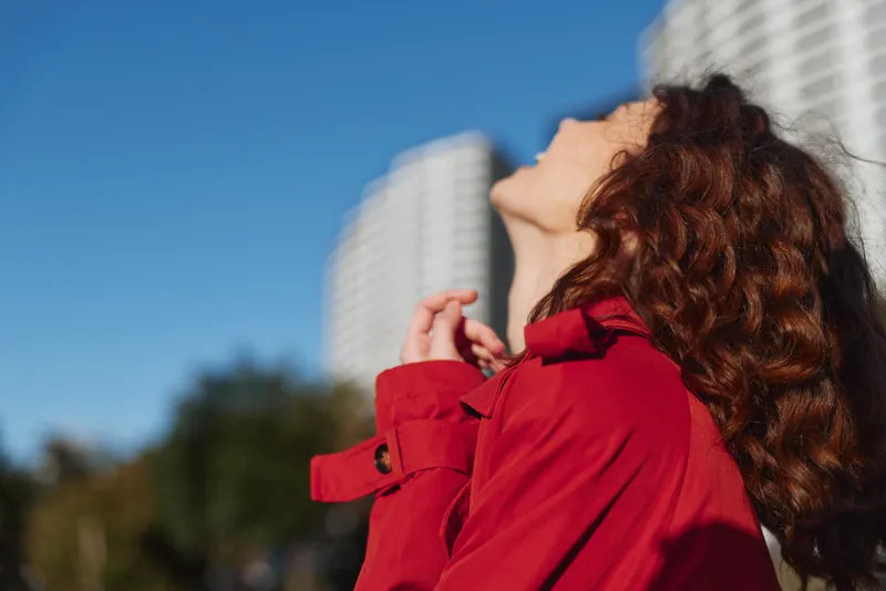 A woman in a red coat gazes upward with awe in a sunlit urban plaza, tall buildings rising behind her, hair catching the light for a moment of possibility.
