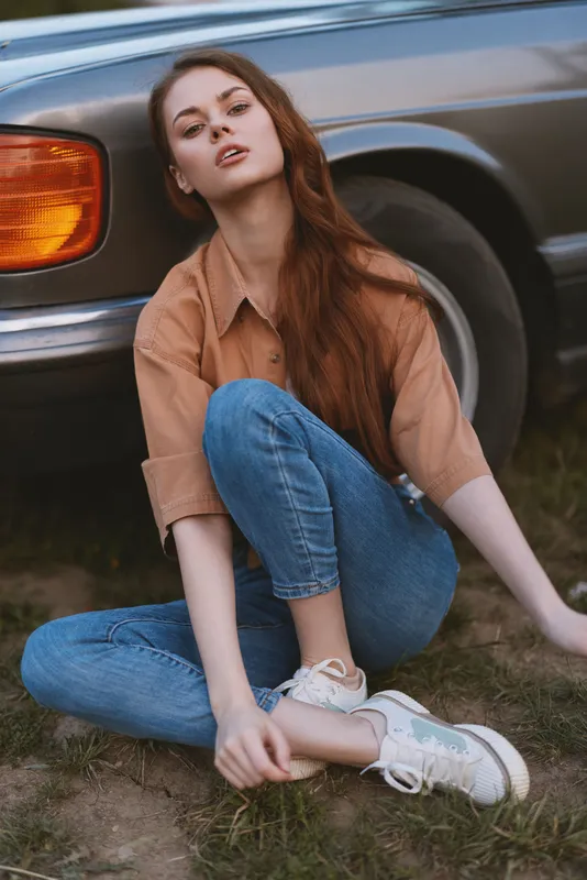 Young woman sitting casually in jeans and a brown shirt by a classic car, expressing relaxed confidence in a natural outdoor setting.