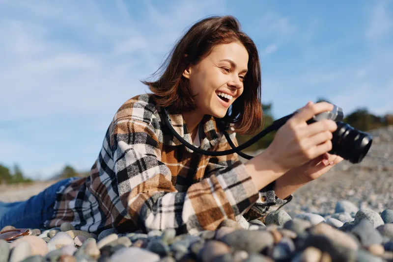 Photographer lying on a rocky beach, smiling while shooting with a camera, enjoying outdoor photography and a relaxed lifestyle in bright sunlight and natural scenery.