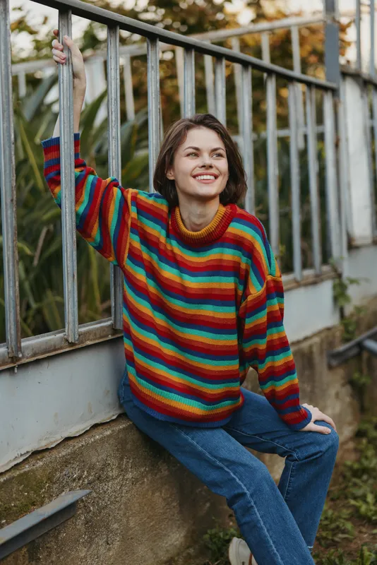 Woman in colorful striped sweater smile in casual portrait outdoors, sitting by metal railing with jeans, relaxed young adult enjoying urban garden atmosphere and natural light.