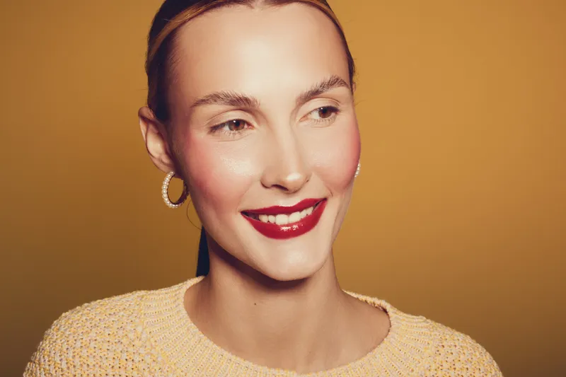 Smiling woman with red lipstick and hoop earrings looks away on warm brown background. Close-up portrait with natural makeup and cozy sweater. Concept of beauty, fashion, and happiness.