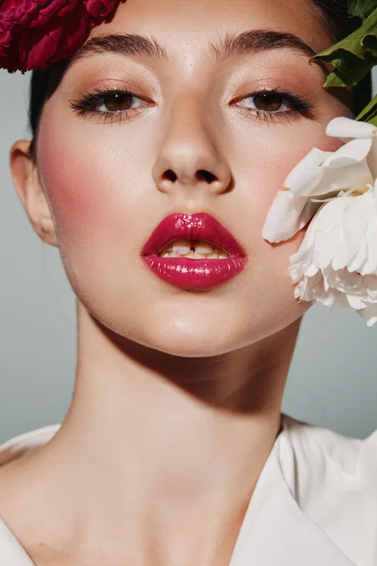 Close-up of a woman with glossy red lips and glowing skin posing with flowers by her face. Beauty, creative makeup, fashion styling on light background.