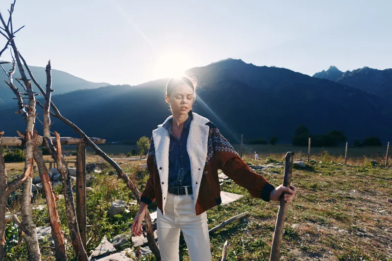 Woman in shearling jacket stands outdoors by rustic fence with walking stick, backlit by bright sun and dramatic mountains, rural landscape, contemplative mood and natural light.