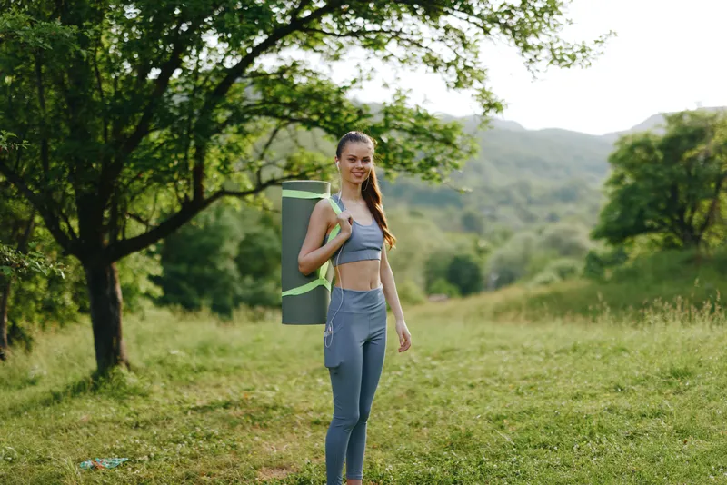 Young woman in athletic wear smiling with yoga mat, standing in a lush green landscape, embodying fitness and outdoor wellness in summer