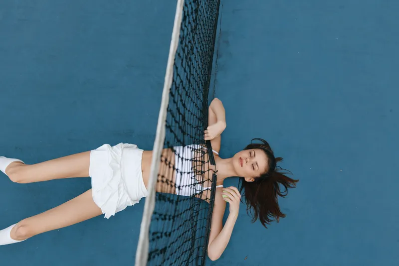 A young woman in sporty attire relaxing on a tennis court, showing a blend of leisure and athleticism against a vibrant blue background