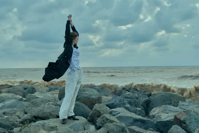 Woman on rocky shore raises arms in windy weather, cape fluttering, wearing white trousers and dark jacket, capturing freedom and resilience beside the sea with dynamic outdoor motion, dramatic mood