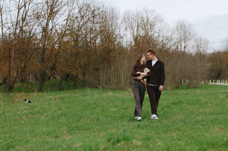 A couple walks through a grassy field with a young goat kid, sharing a gentle moment. Casual clothing, soft light, and open space convey rural lifestyle and connection.